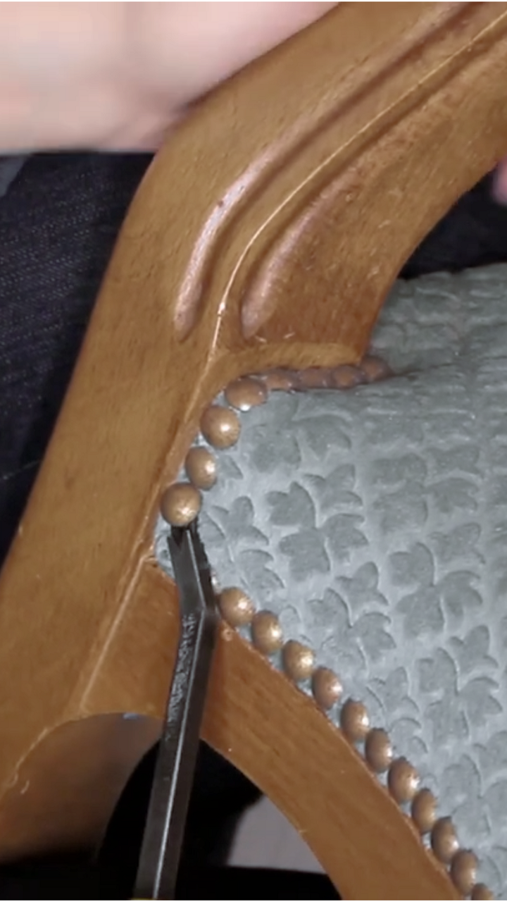 Gros plan sur l’utilisation d’un arrache-agrafes pour retirer des clous d’un fauteuil Voltaire pendant la phase de dégarnissage. Explication des bonnes pratiques pour éviter d’abîmer le bois et des précautions de sécurité pour ne pas se blesser.