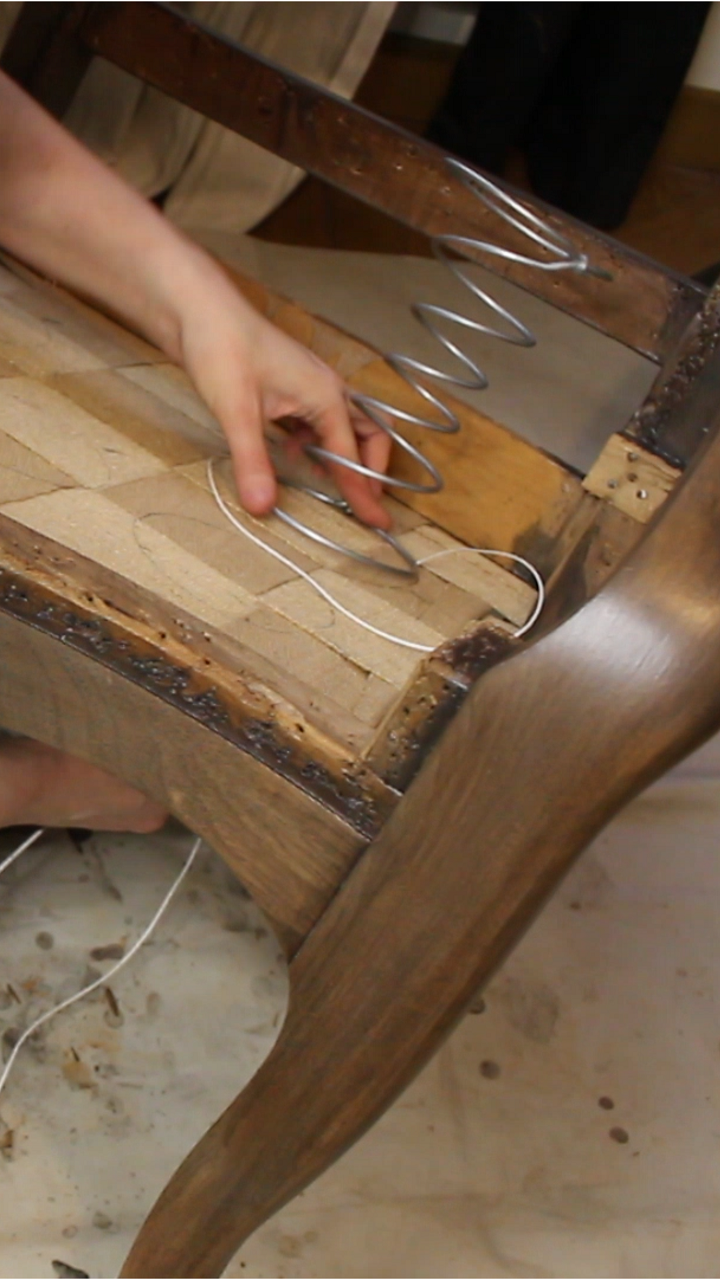 Pose d'un ressort sur un sanglage en jute de sur l’assise d’un fauteuil Voltaire en cours de restauration. Explication de l’importance du sanglage pour assurer la solidité du siège.