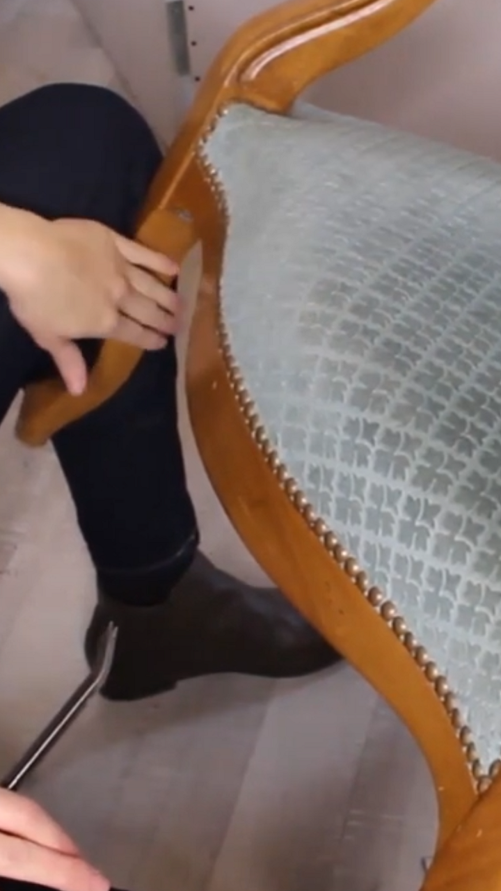 Tapissier en train de dégarnir un fauteuil Voltaire avec les pieds du fauteuil bloqués entre les jambes. Explication des différentes positions de travail en tapisserie pour optimiser le confort et éviter les douleurs dorsales.